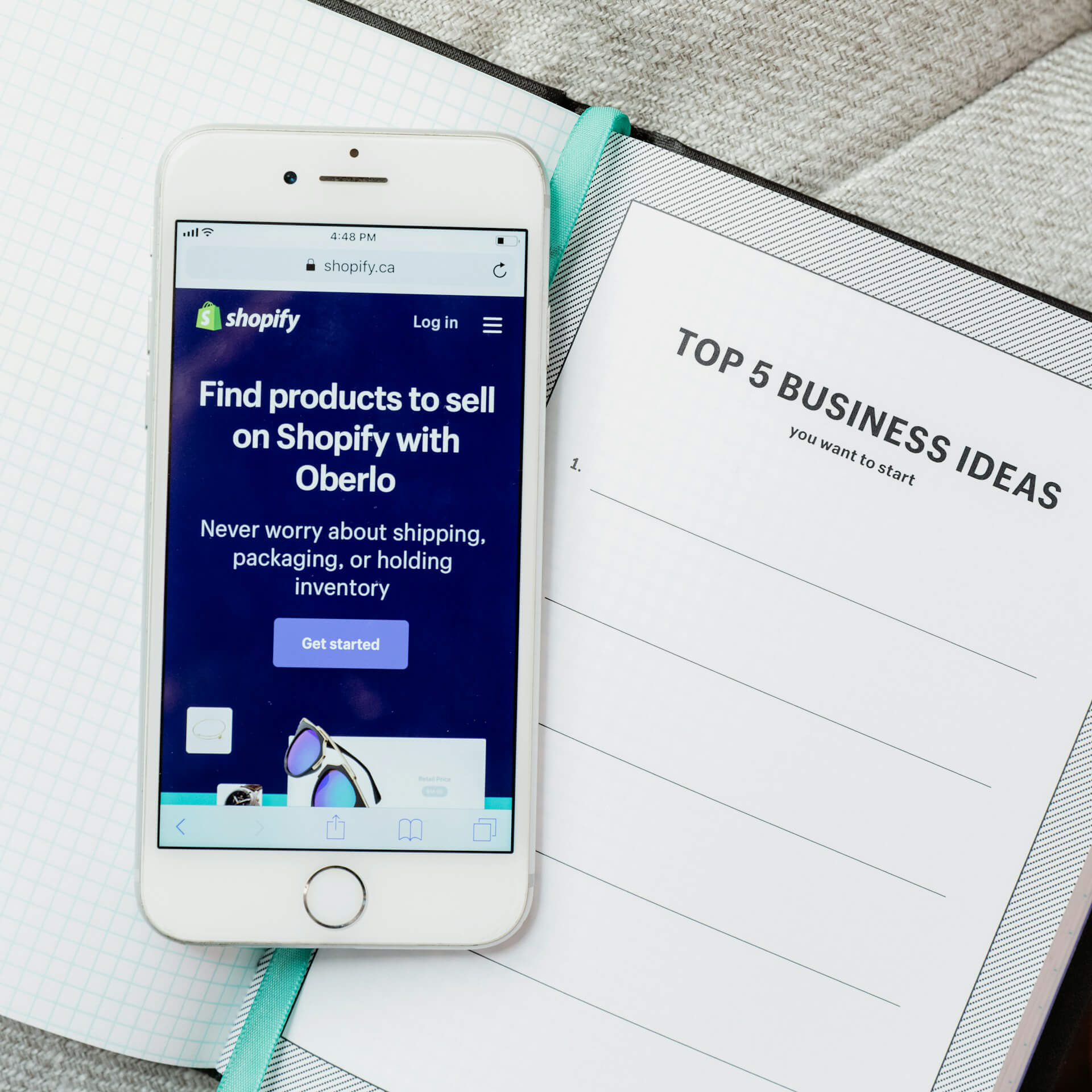 iPhone displaying Shopify's Oberlo page with the headline 'Find products to sell on Shopify', placed on a business planner titled 'Top 5 Business Ideas', ideal for entrepreneurs starting an eCommerce store.